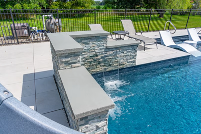 Three-tiered stacked stone waterfall cascading into a modern pool with lush green surroundings