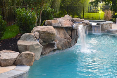Multi-tiered natural stone boulder waterfalls cascading into a swimming pool with lush garden greenery