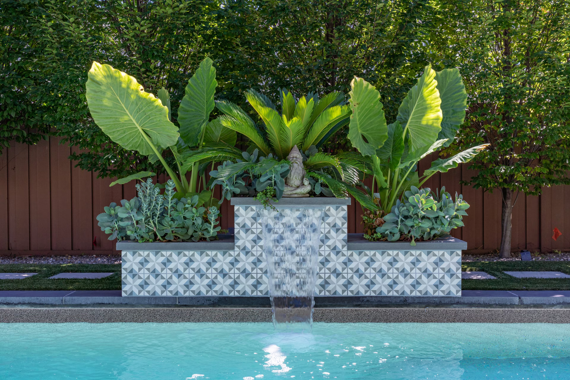 Three-tiered geometric tiled waterfall spilling into a pool surrounded by lush tropical greenery