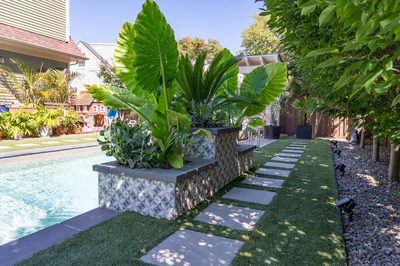 Multi-tiered tiled planter with tropical greenery beside a swimming pool and stone walkway