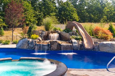 Two-tiered natural stone boulder waterfall cascading into a pool surrounded by lush trees and plants