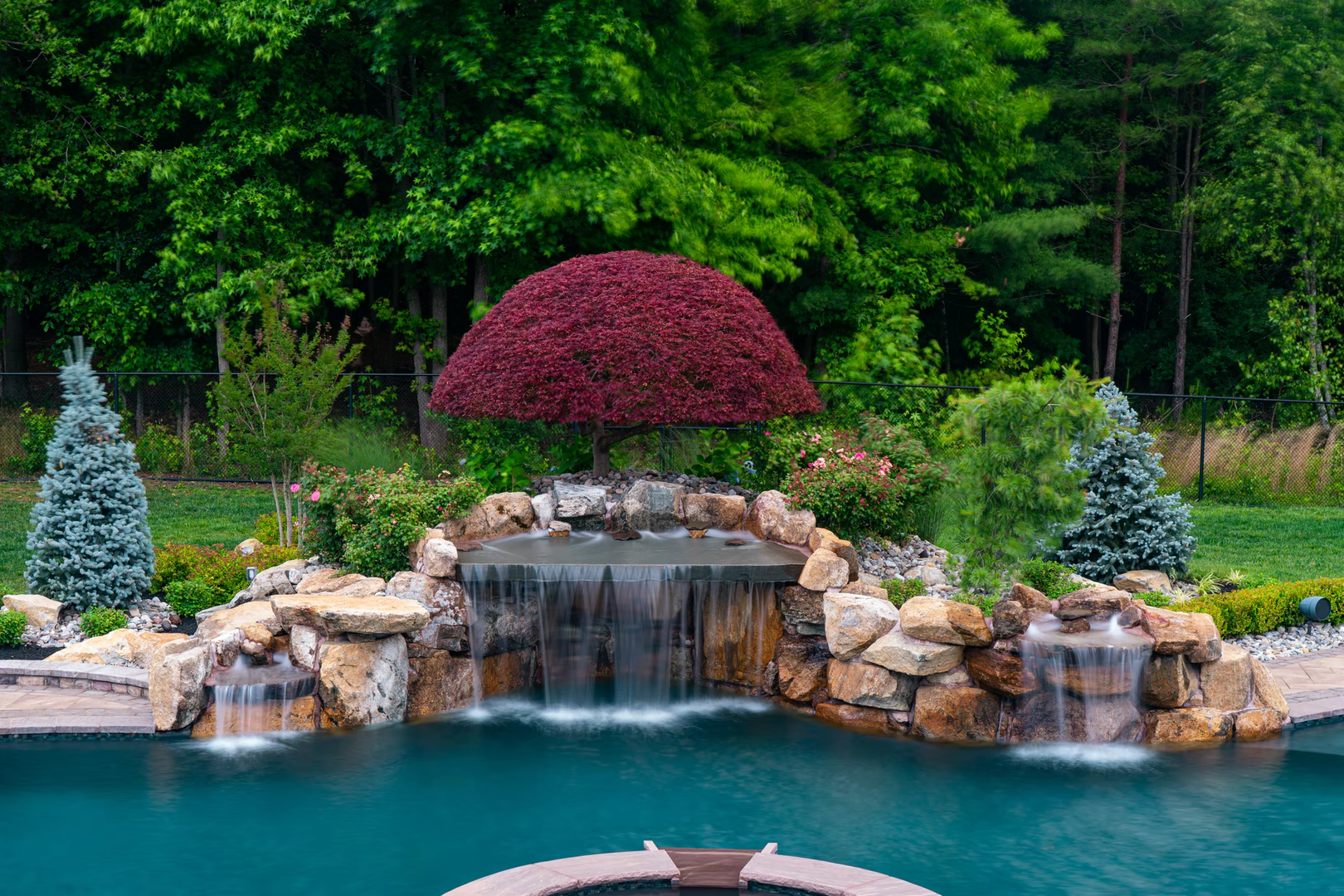 Boulder waterfall cascading into a pool with lush greenery and a red maple