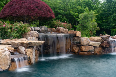 Multi-tiered natural stone boulder waterfalls cascading into a pool surrounded by lush garden plants