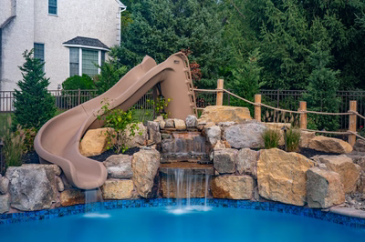 Two-tiered natural stone boulder waterfall cascading into a pool with lush landscaping and slide