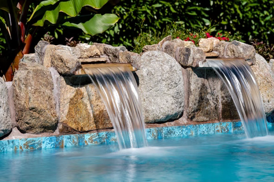 Dual sheet-flow natural stone waterfalls cascading into a tiled pool bordered by lush tropical plants