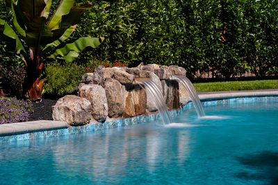 Twin sheet waterfalls cascading from natural stone boulders into a pool with lush tropical plants