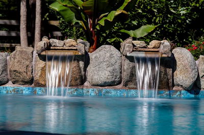 Natural stone boulder waterfalls spill into a tiled pool surrounded by lush tropical plants