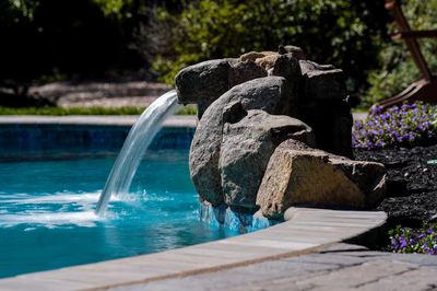 Natural stone boulder waterfall cascading into a blue pool surrounded by lush garden plants