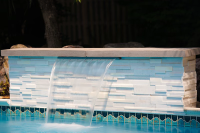 Single-tier sheer descent waterfall cascading from white stone wall into pool with background foliage