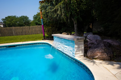 Single-tier sheer descent waterfall cascading into a backyard pool bordered by boulders and trees