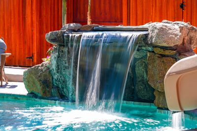 Single-tier natural stone boulder waterfall cascading into a pool with nearby plants and fencing