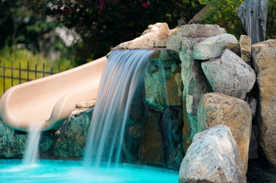 Multi-tiered natural stone boulder waterfall cascading into a swimming pool with surrounding garden plants