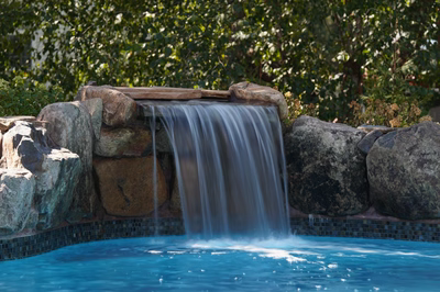 Single-tier natural stone boulder waterfall cascading into a blue pool with lush green foliage