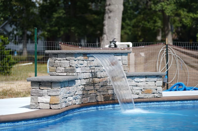 Three-tiered stone waterfall with sheer flow cascading into a pool near trees and fencing