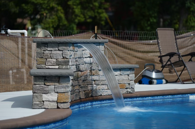 Two-tiered natural stone boulder waterfall cascading into a swimming pool surrounded by lush green trees