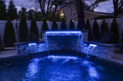Triple-tiered natural stone boulder waterfall with blue lighting cascading into a pool near evergreens