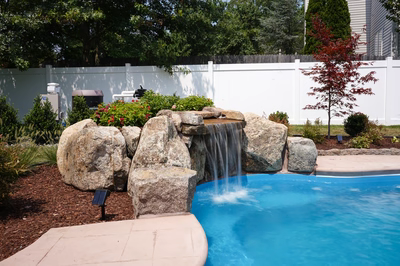Single-tier natural stone boulder waterfall cascading into a blue pool with lush garden plants