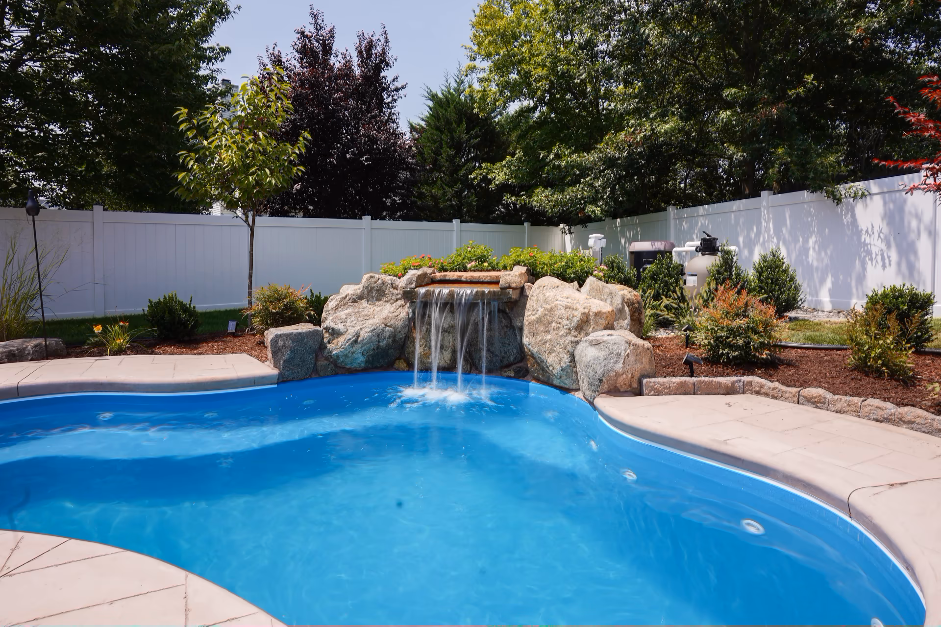 Single-tier natural stone boulder waterfall cascading into a blue pool with surrounding garden plants