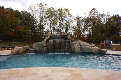 Two-tiered natural stone boulder waterfall cascading into a swimming pool framed by autumn trees