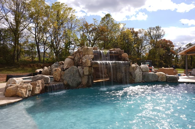 Multi-tiered natural stone boulder waterfall cascading into a swimming pool surrounded by lush trees