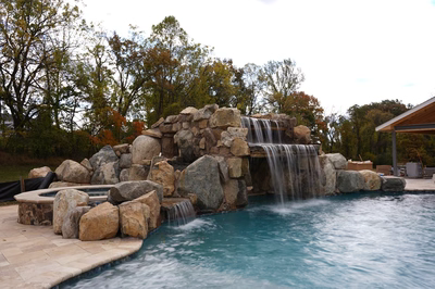 Multi-tiered natural stone boulder waterfall cascading into a pool surrounded by tall deciduous trees