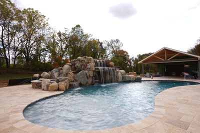 Two-tiered natural stone boulder waterfall cascading into a freeform pool with surrounding autumn trees